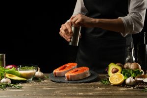 The chef prepares fresh salmon fish, smorgu trout, sprinkling black pepper with the ingredients. Frost in the air. cooking fish food. Salmon steak. Cooking vegan cuisine, restaurants, hotel business