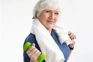 Strength, energy, wellness and healthy active lifestyle concept. Stylish athletic senior female with fit body and gray hair wrking out in gym using dumbbell, wearing white towel around her neck