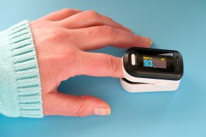 Pulse Oximeter portable digital device to measure persons oxygen saturation. Reduction in oxygenation is an emergency sign of Covid-19 viral pneumonia. Device on female hand, blue mint background.