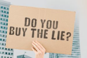 The question " Do you buy the lie? " on a banner in hand with blurred background. False. Liar. Lies. Statement. Fraud. Service. Commerce. Selling. Internet. Money. Loss