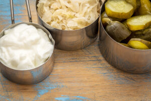 sauerkraut, cucumber pickles and yogurt - popular probiotic fermented food - three measuring cups against rustic wood